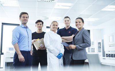 Five people stand together in a modern laboratory; one wears a lab coat and gloves, another holds a box, and two hold documents.