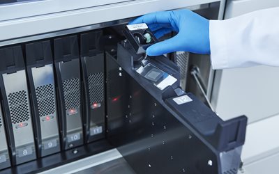 A gloved hand places a sample tray into a laboratory sequencing machine with multiple numbered slots.