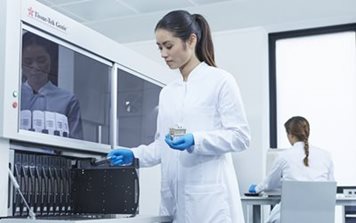 A woman in a lab coat and gloves operates laboratory equipment while another person works at a desk in the background.