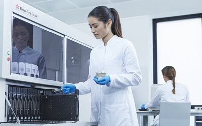 A woman in a lab coat and gloves operates scientific equipment, while another person works at a desk in the background of a laboratory.