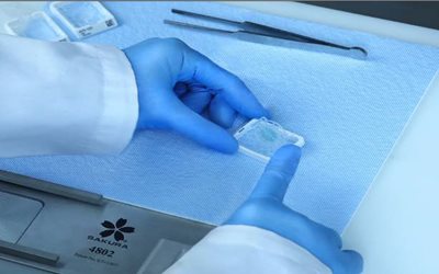 A person wearing blue gloves handles a microscope slide on a lab mat, with forceps and sample containers nearby.