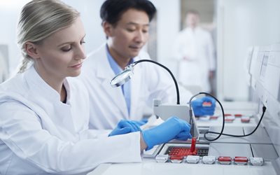 Two scientists in lab coats work with laboratory equipment, handling samples and using a magnifying device in a modern laboratory setting.