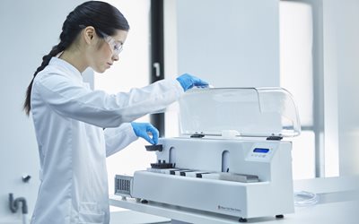 A person in a lab coat and gloves operates a laboratory instrument, placing a sample into the machine in a bright, modern lab setting.