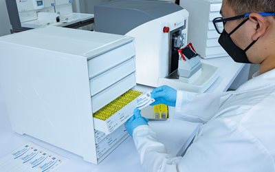 A lab technician wearing gloves and a mask retrieves a labeled sample from a drawer in a laboratory setting.