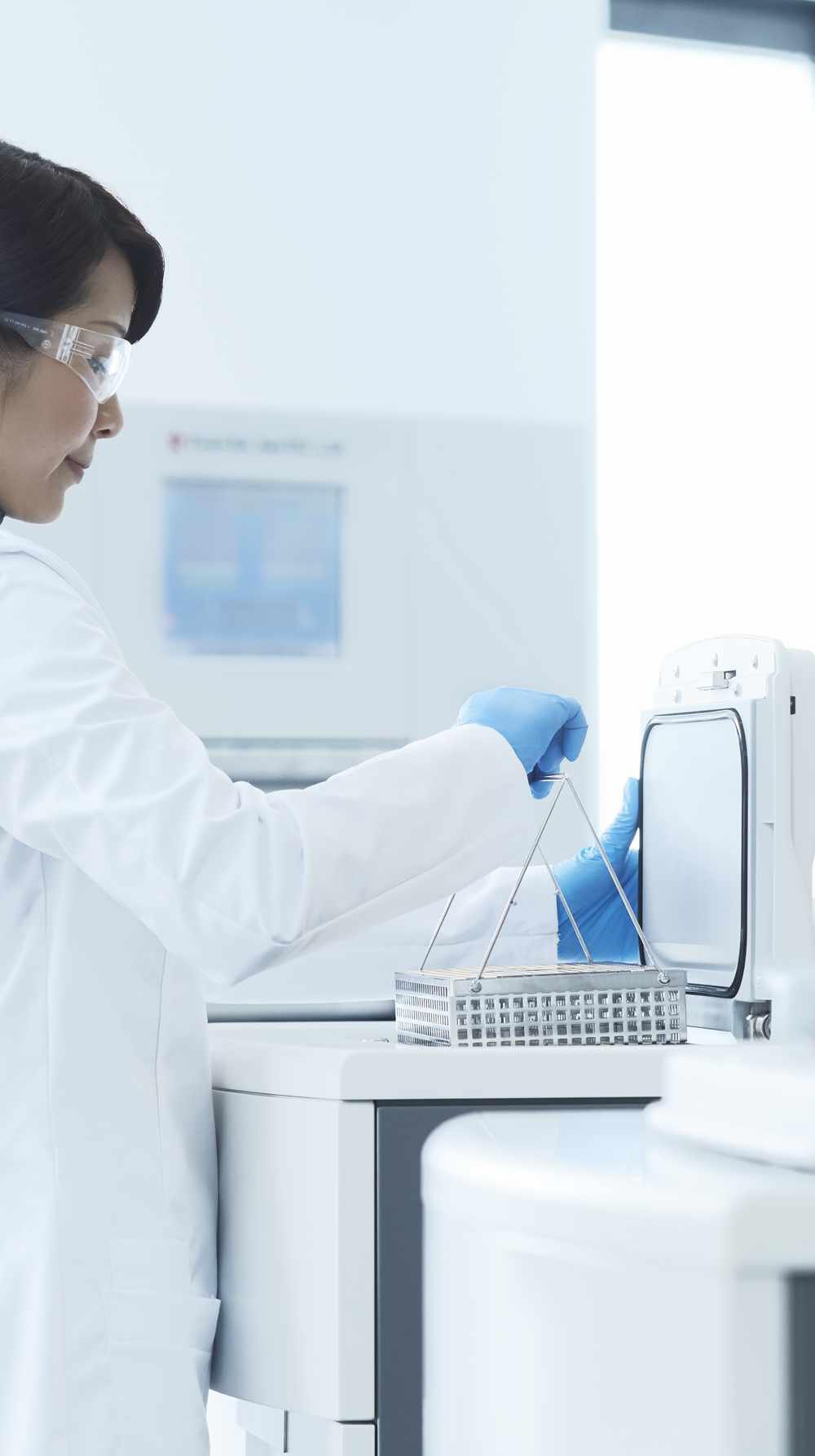 A scientist wearing a lab coat and gloves handles a tray of test tubes in a modern laboratory setting.