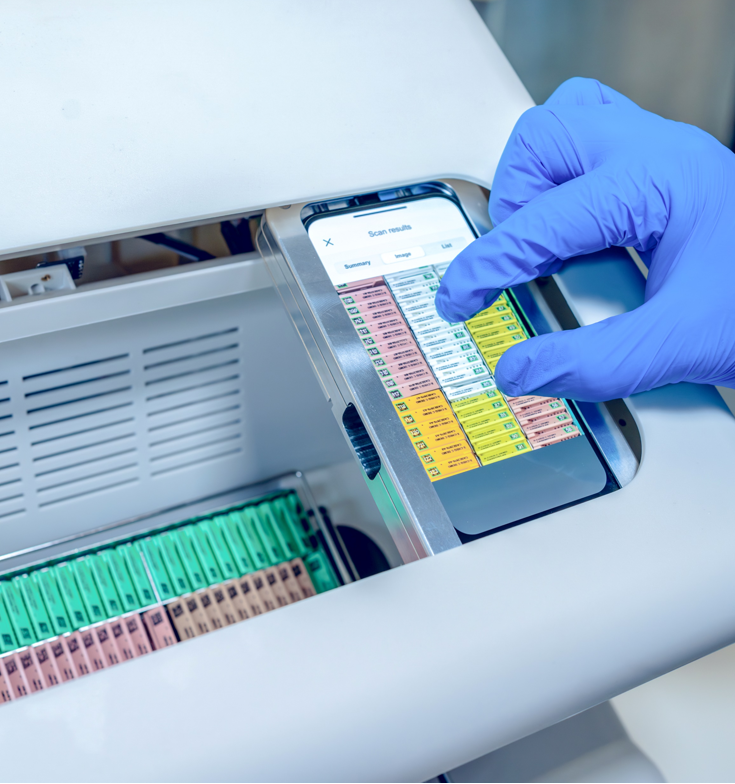 A gloved hand uses a touchscreen device to scan color-coded slides in a laboratory setting.