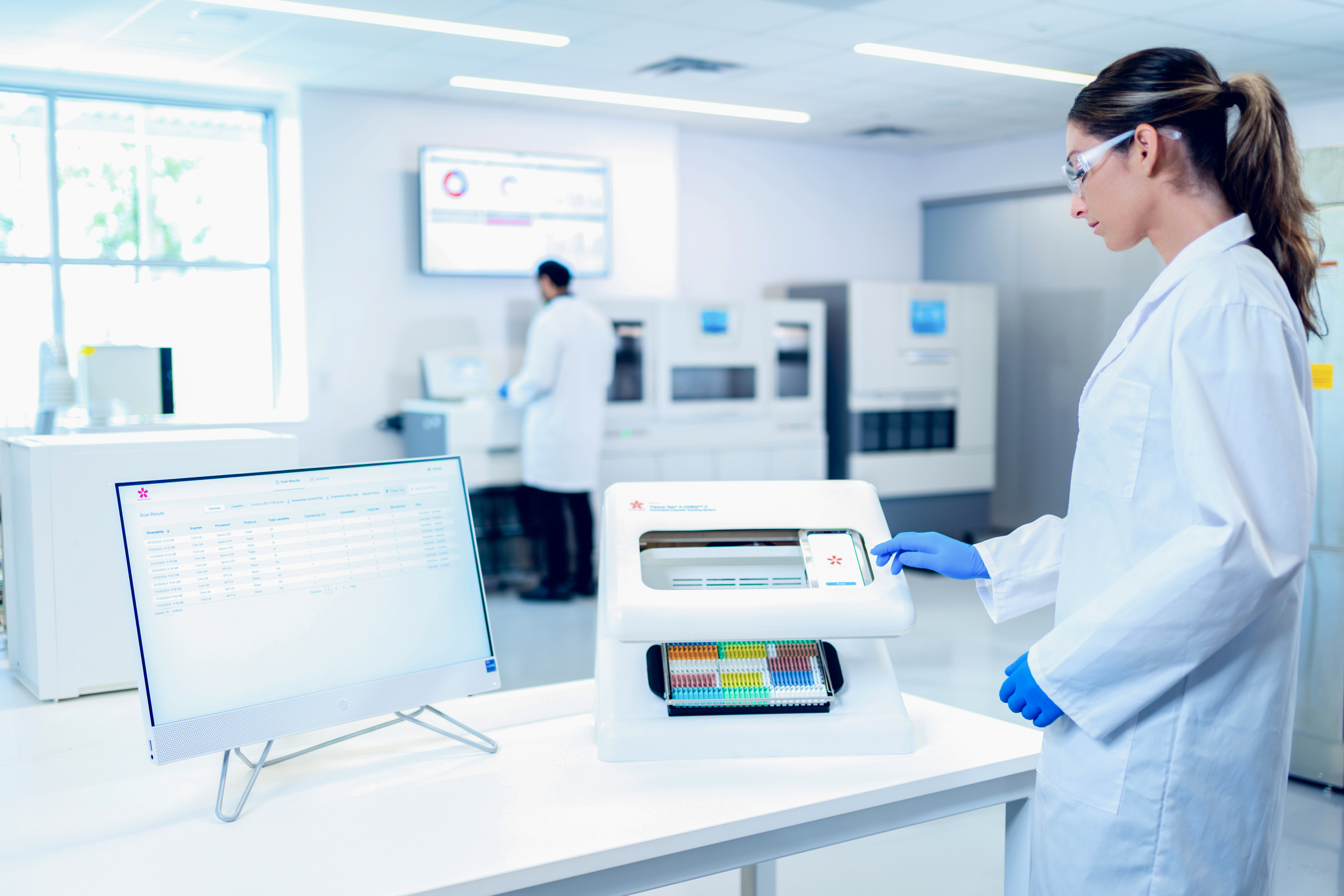 A lab technician operates an automated analyzer with test tubes, while another person works at a computer in a bright laboratory.