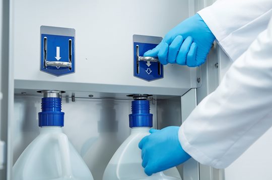 Person wearing blue gloves installs large chemical containers into a dispensing system with labeled slots inside a laboratory or industrial setting.