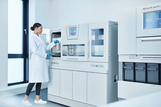 A lab technician in a white coat uses a large automated analyzer in a modern, bright laboratory setting.