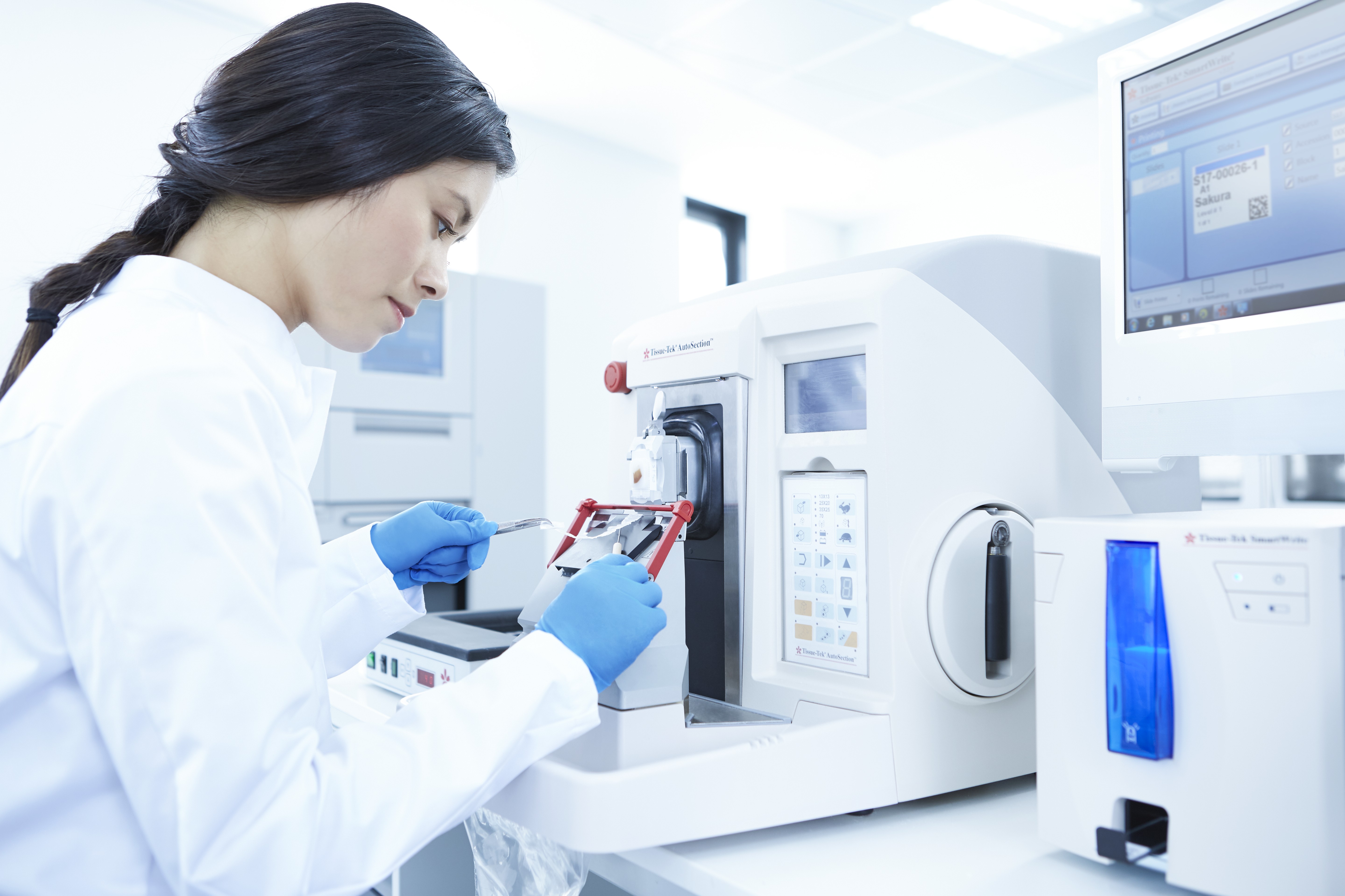 A scientist in a lab coat and gloves operates a microtome machine, preparing samples on slides with a computer monitor visible nearby.