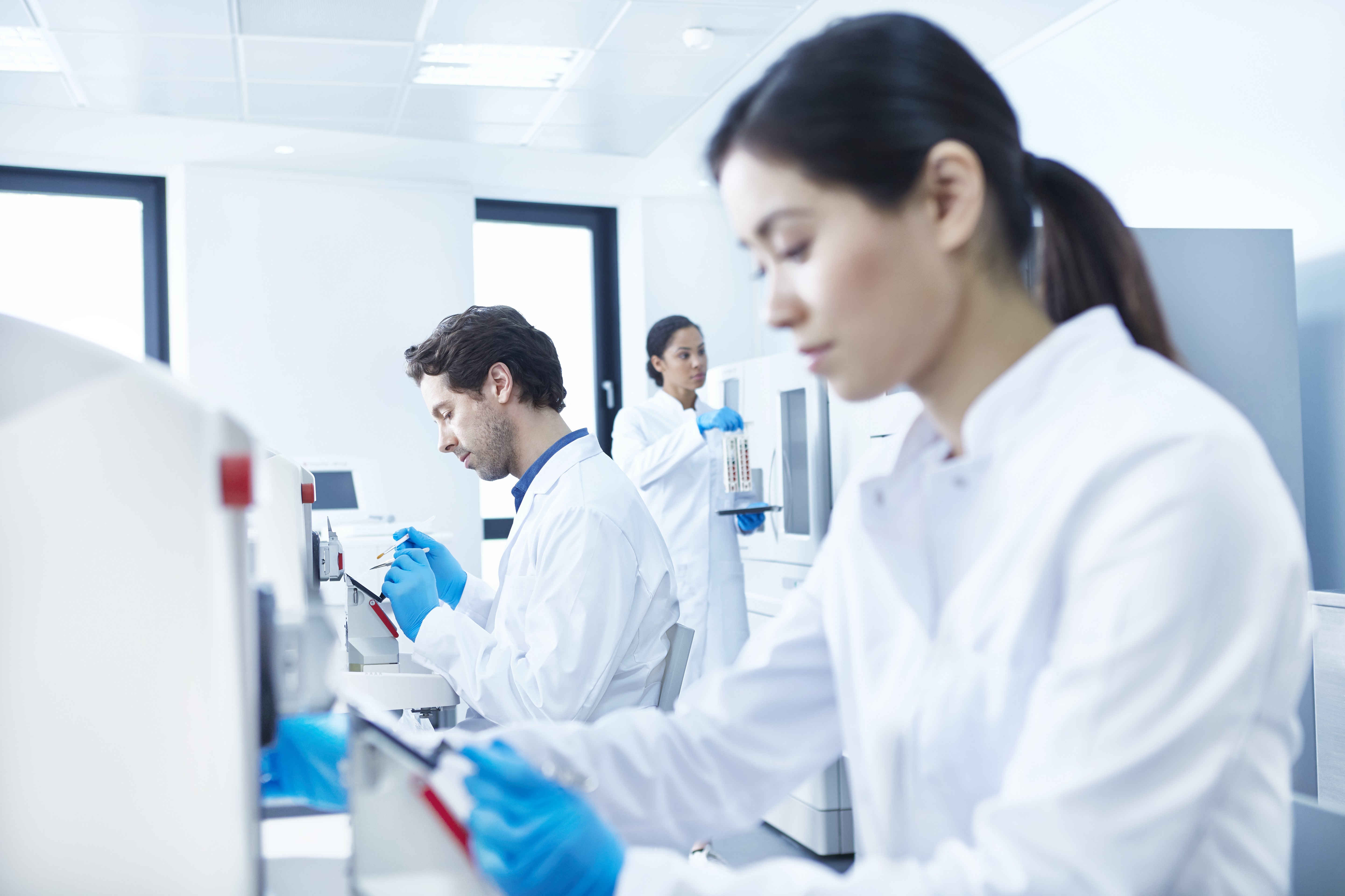 Three scientists in lab coats and gloves work with laboratory equipment and samples in a bright, modern laboratory setting.