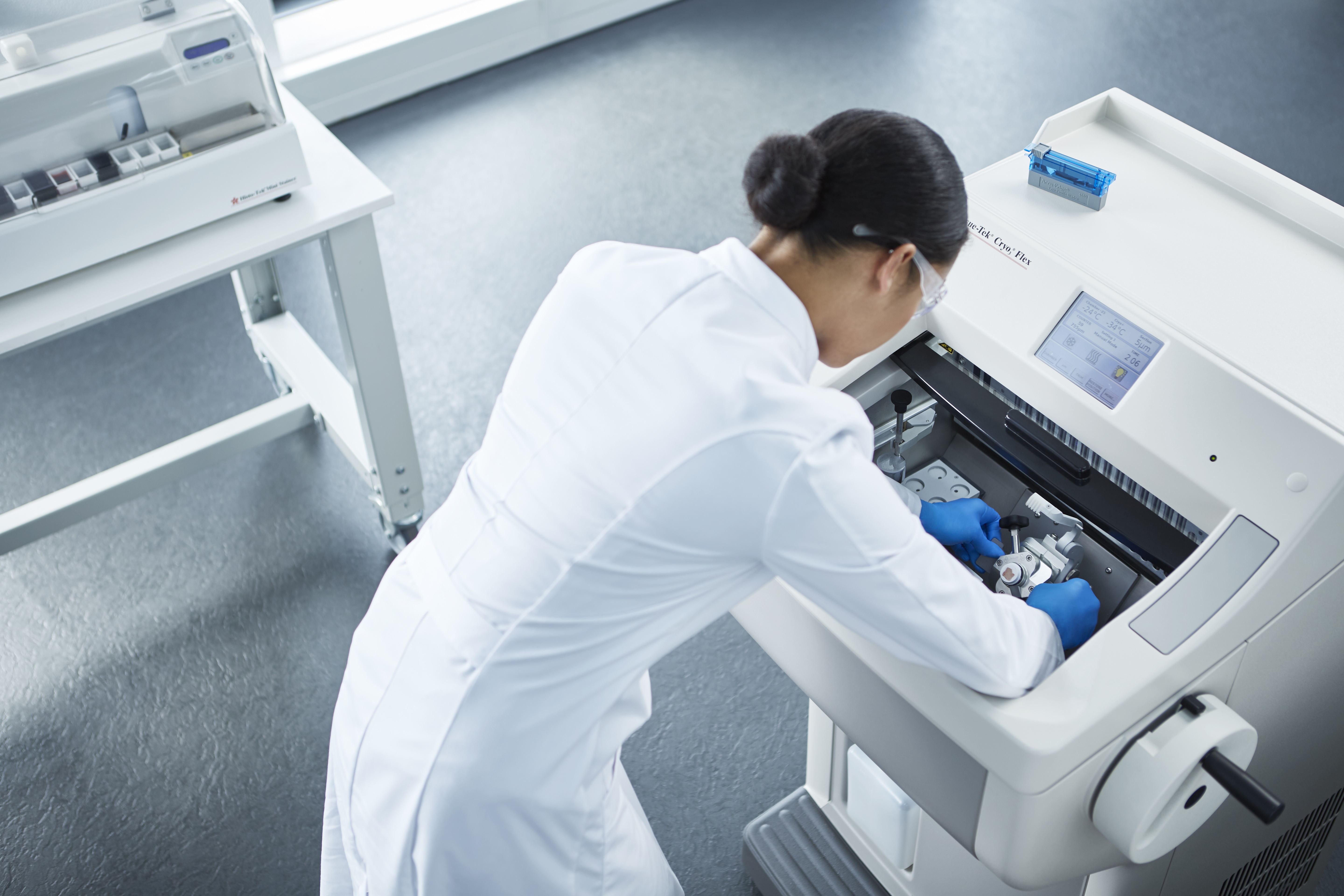 A scientist in a white lab coat and gloves operates a laboratory machine, adjusting equipment inside a compartment in a clinical setting.