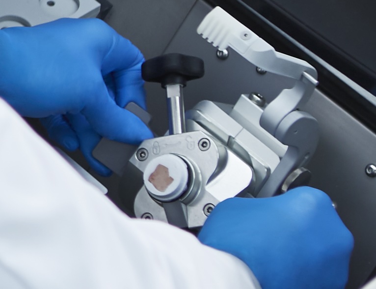 A person wearing blue gloves operates a laboratory microtome with a sample block mounted in the device.