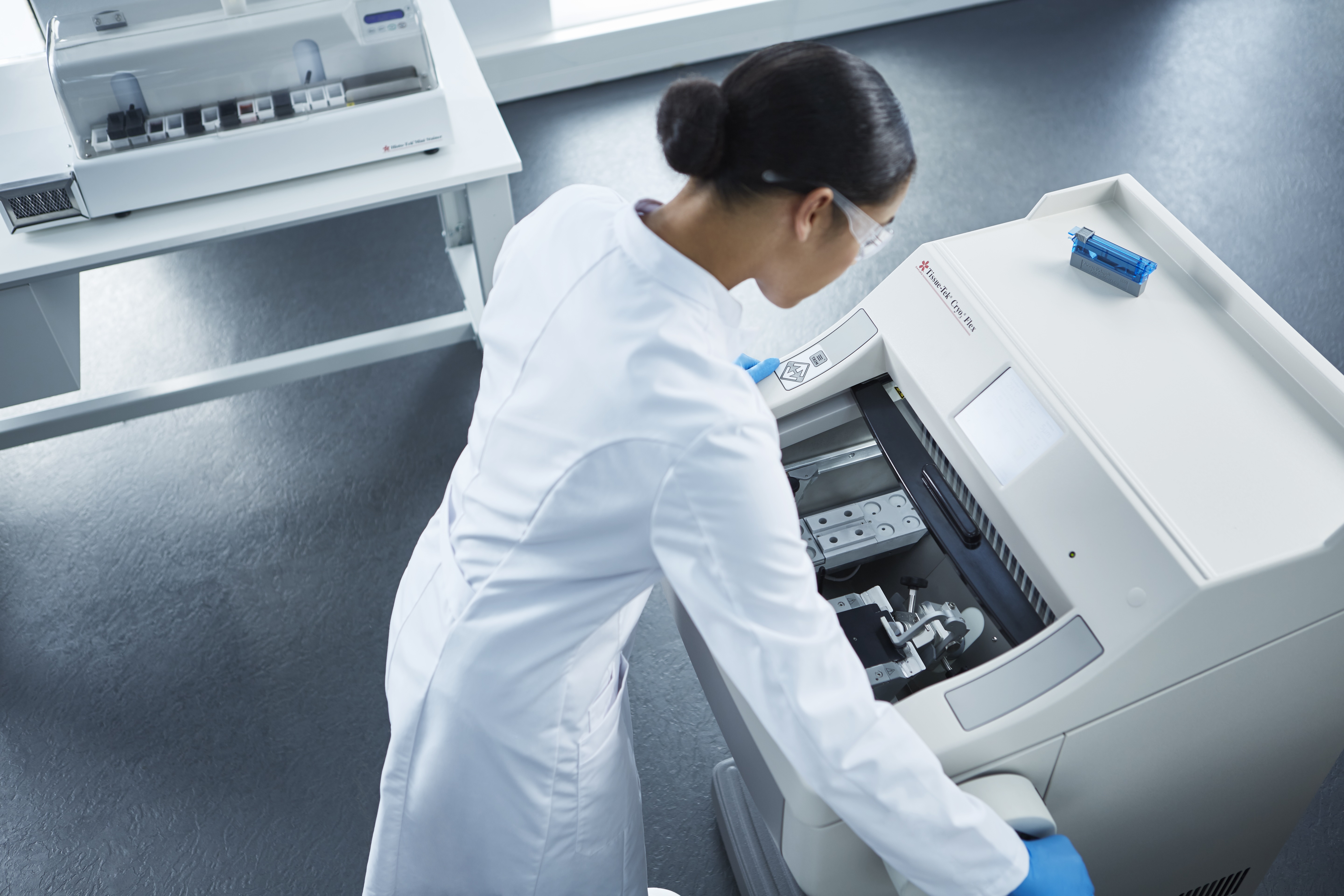 A person in a white lab coat and safety glasses operates a laboratory analyzer machine in a clinical lab setting.