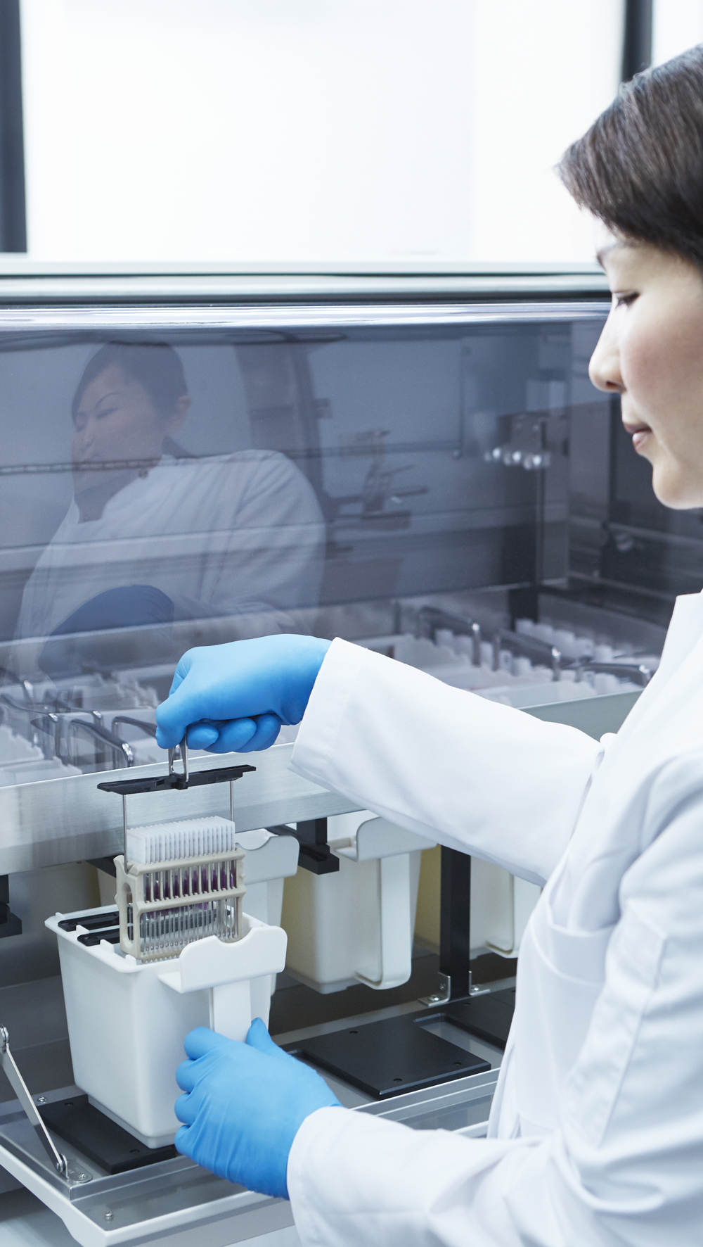 A scientist in a lab coat and blue gloves operates laboratory equipment, handling a tray with multiple test tubes inside a sterile enclosure.