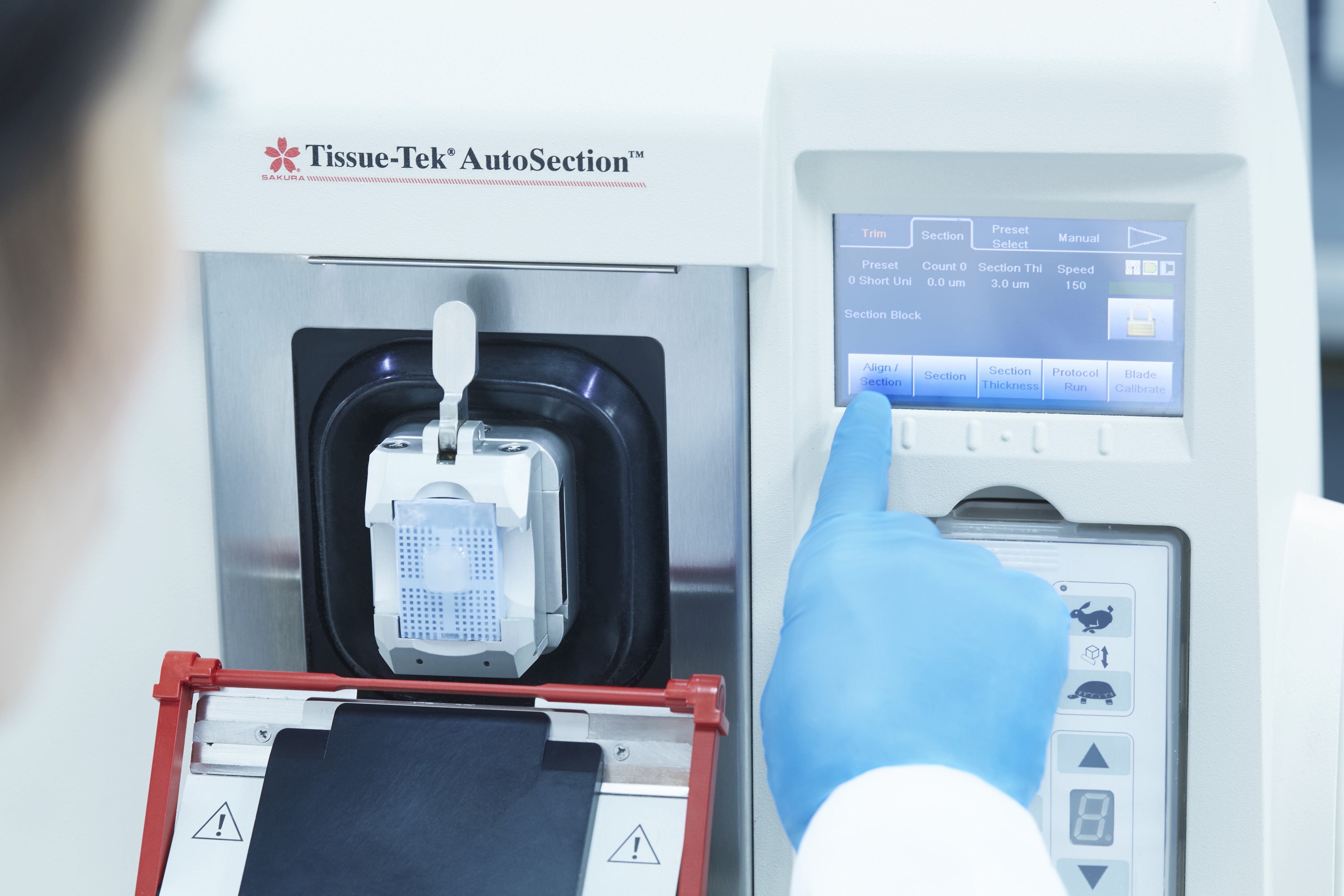 A gloved person operates a Tissue-Tek AutoSection machine, pressing a button on the touchscreen to prepare a sample for sectioning.