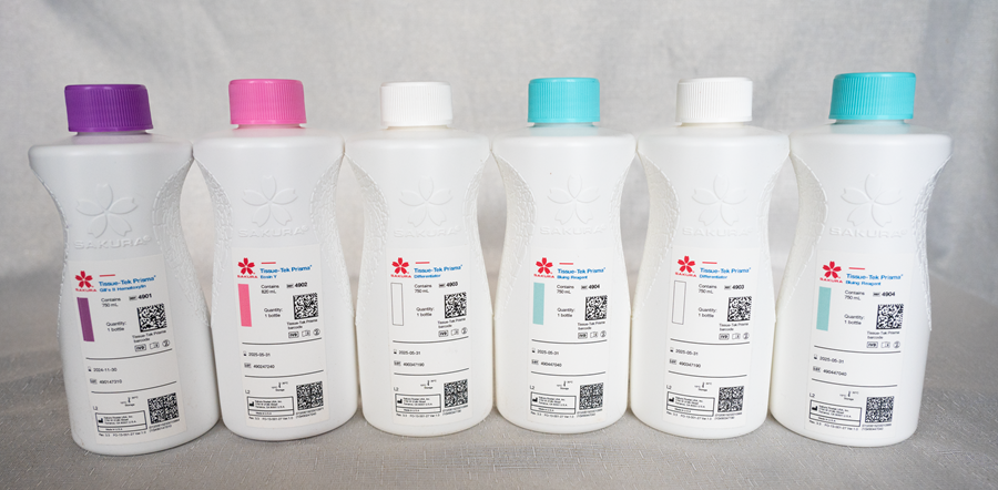 Six white plastic bottles with colored caps (purple, pink, blue, turquoise) labeled as Sakura tissue ink reagent are lined up against a plain background.