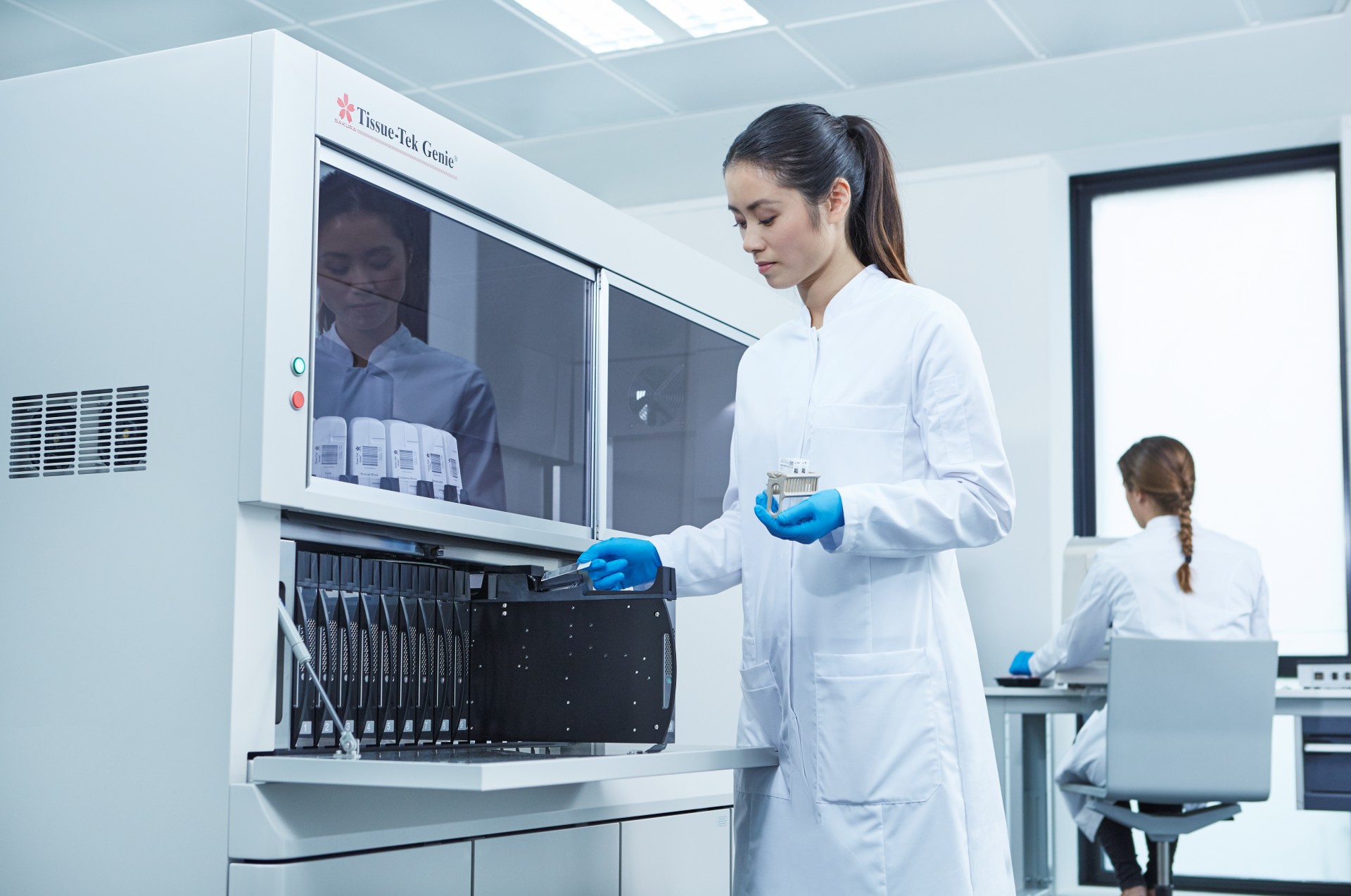 Two scientists in lab coats work in a modern laboratory; one operates equipment with vials, while the other sits at a computer in the background.